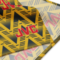 A close-up of a JVC-branded textile product, likely a towel or blanket, featuring a bold geometric pattern in yellow, navy, and gray with large red JVC logos.