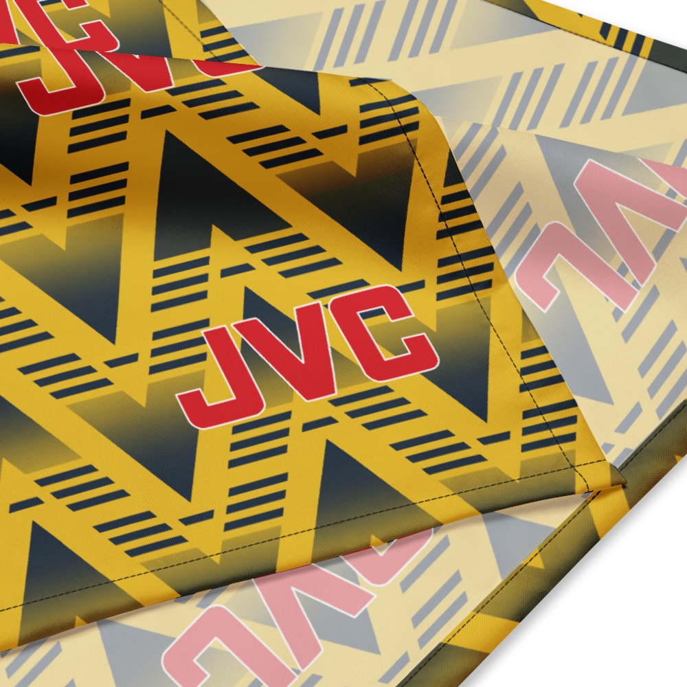 A close-up of a JVC-branded textile product, likely a towel or blanket, featuring a bold geometric pattern in yellow, navy, and gray with large red JVC logos.