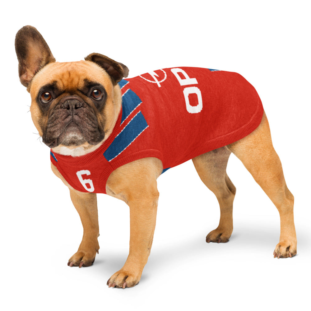 A fawn French bulldog wearing a red knit dog sweater with blue and white accents, standing against a plain white background.