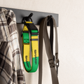 A close-up of a black wall-mounted hook rack holding two straps: a bright green and yellow strap with black hardware and the word SHARP printed vertically, and a taupe-gray strap hanging beside it. A plaid shirt is partially visible on the left, and the wall occupies the background.