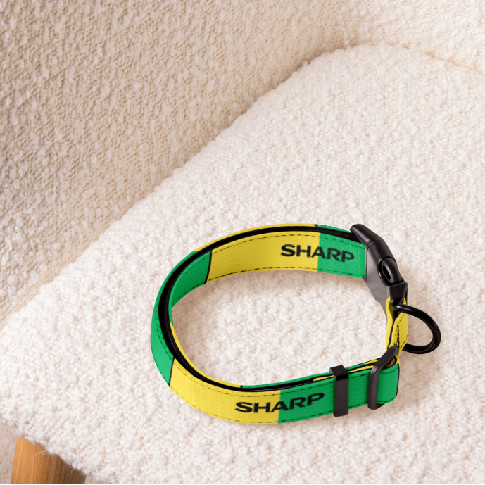 A pet collar with a yellow and green checkered pattern and the brand name 'SHARP' printed on it, resting on a textured cream boucle chair.