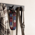 A dog collar and a long leash hang from a black wall hook on a light textured wall. The dog collar has a patchwork design in burgundy, blue, and gray with circular star details, while the leash is a solid taupe-brown color.