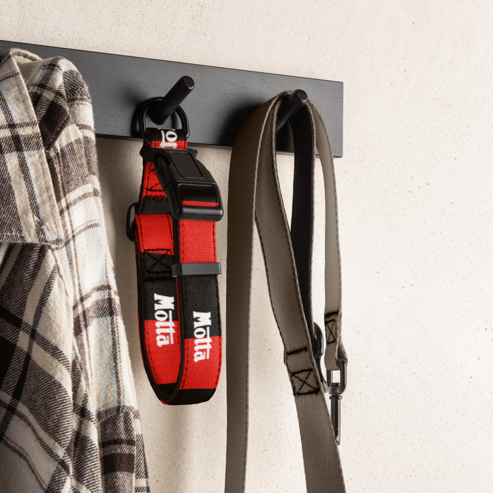 A red and black Motta dog collar hangs on a black wall hook next to a gray strap, with a plaid shirt partially visible on the left, against a light textured wall.
