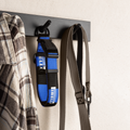 A blue and black IRELI dog collar hangs on a black wall hook next to a plaid shirt, with a gray strap hanging on the right, against a light textured wall.