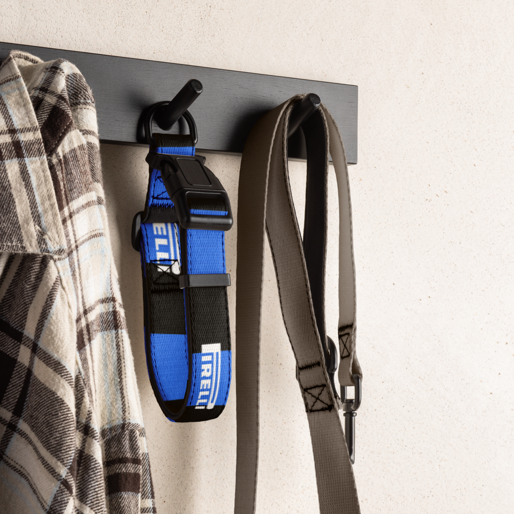 A blue and black IRELI dog collar hangs on a black wall hook next to a plaid shirt, with a gray strap hanging on the right, against a light textured wall.