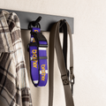 A purple and white dog collar with yellow 'CROOKER' branding hangs on a black wall hook next to a plaid shirt and a gray leash. The background is a textured off-white wall.