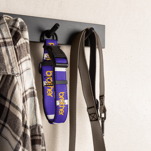 A purple and white dog collar with yellow 'CROOKER' branding hangs on a black wall hook next to a plaid shirt and a gray leash. The background is a textured off-white wall.