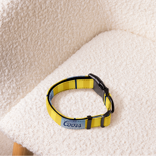 A yellow dog collar with a light blue nameplate reading 'Coors' and black hardware, resting on a cream boucle chair.