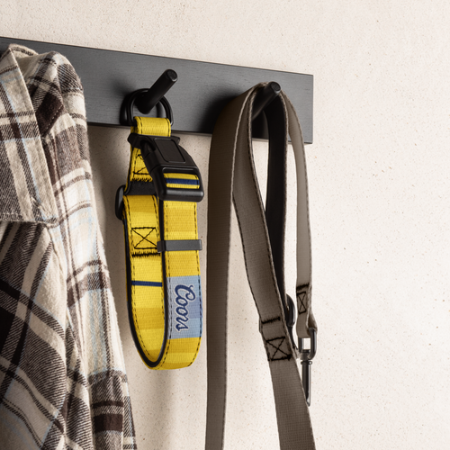 A yellow Coou dog leash with a black plastic buckle hangs on a black wall hook next to a plaid shirt, with a gray strap hanging on the right against a textured off-white wall.