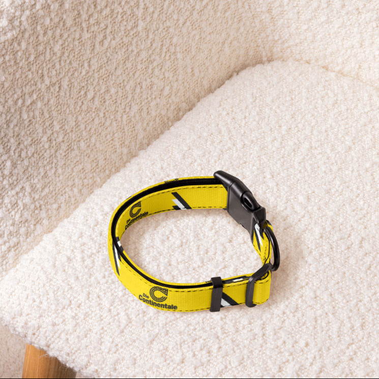 A yellow dog collar with black and white graphic details and a black plastic buckle, resting on a textured cream boucle armchair.