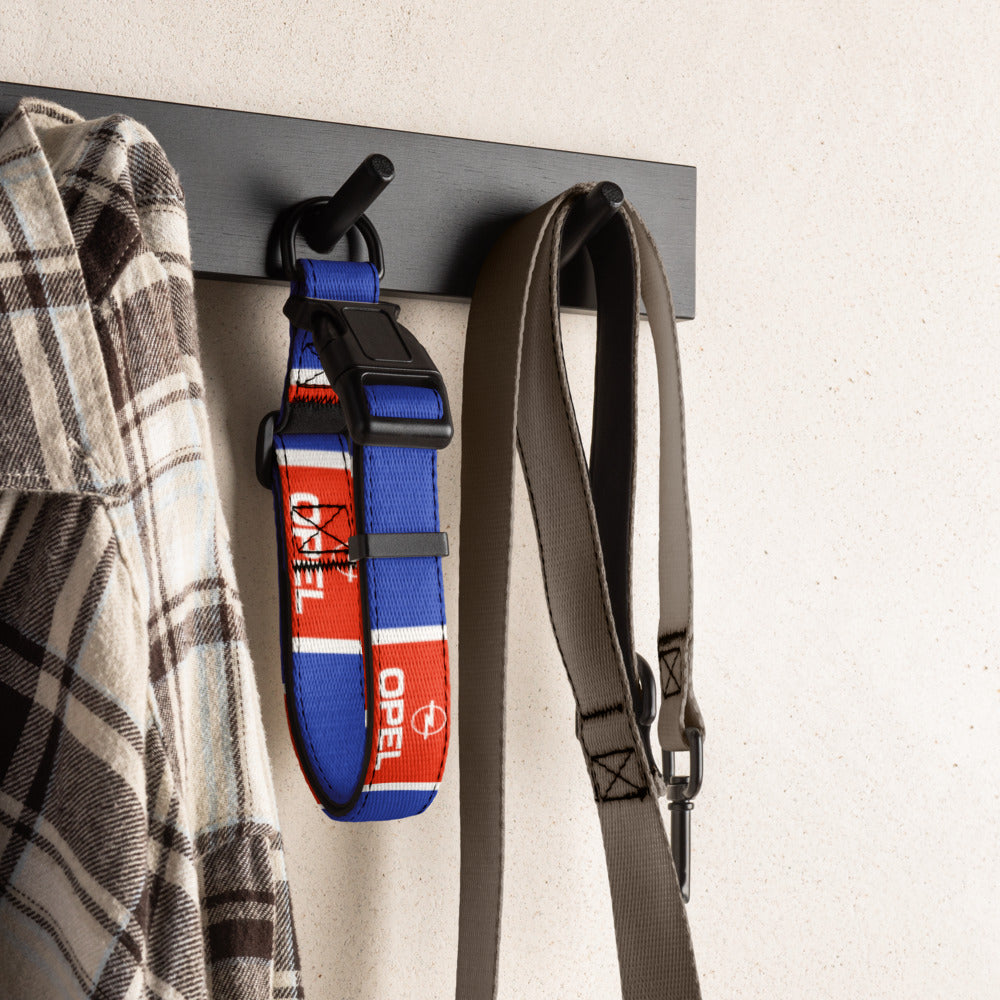 A blue and red dog collar with white 'OPEL' branding hangs on a black wall hook next to a plaid shirt and a gray strap, mounted on a dark horizontal bar against a light textured wall.