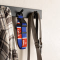 A blue and red dog collar with white 'OPEL' branding hangs on a black wall hook next to a plaid shirt and a gray strap, mounted on a dark horizontal bar against a light textured wall.