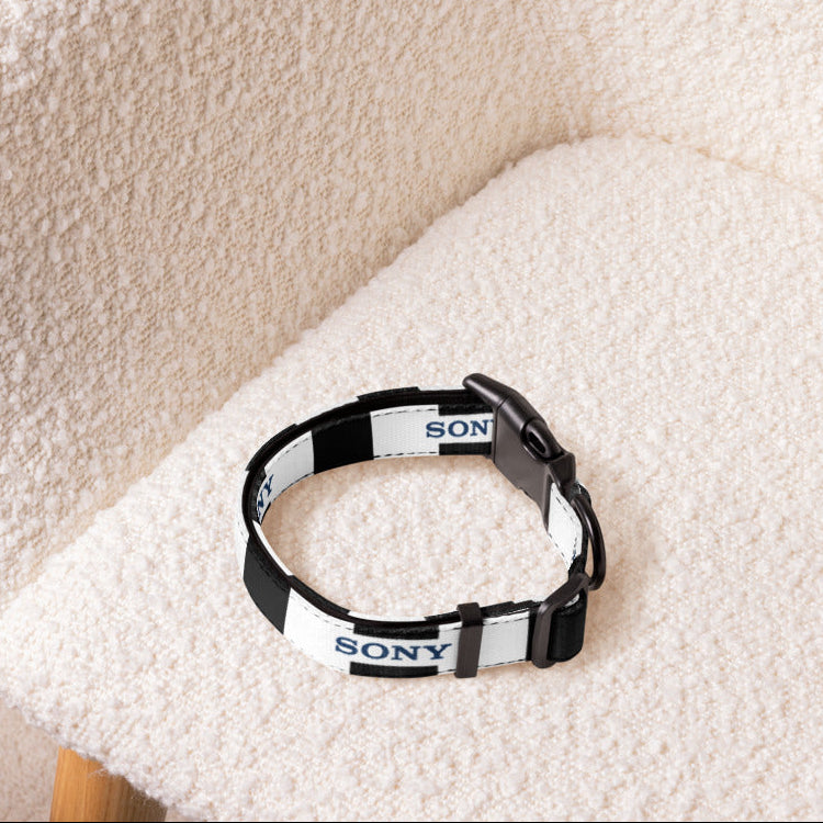 A black and white striped pet collar with 'SONY' printed repeatedly on the strap, resting on a textured cream boucle armchair.